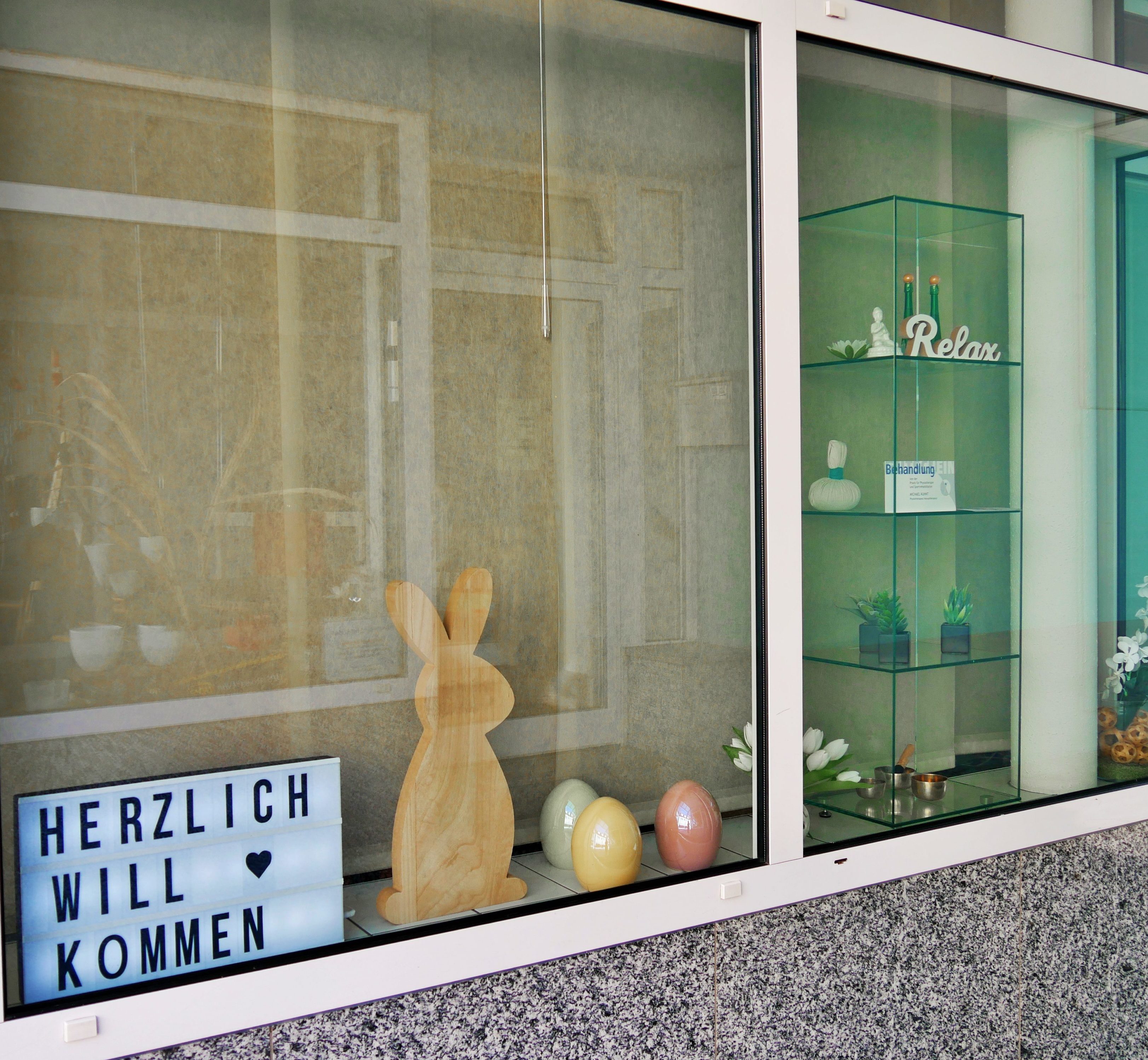 Ein Schaufenster mit einem leuchtenden Schild mit der Beschriftung "Herzlich Willkommen", Oster-Dekoration und einer Glassäule mit verschiedenen Dekoartikeln.