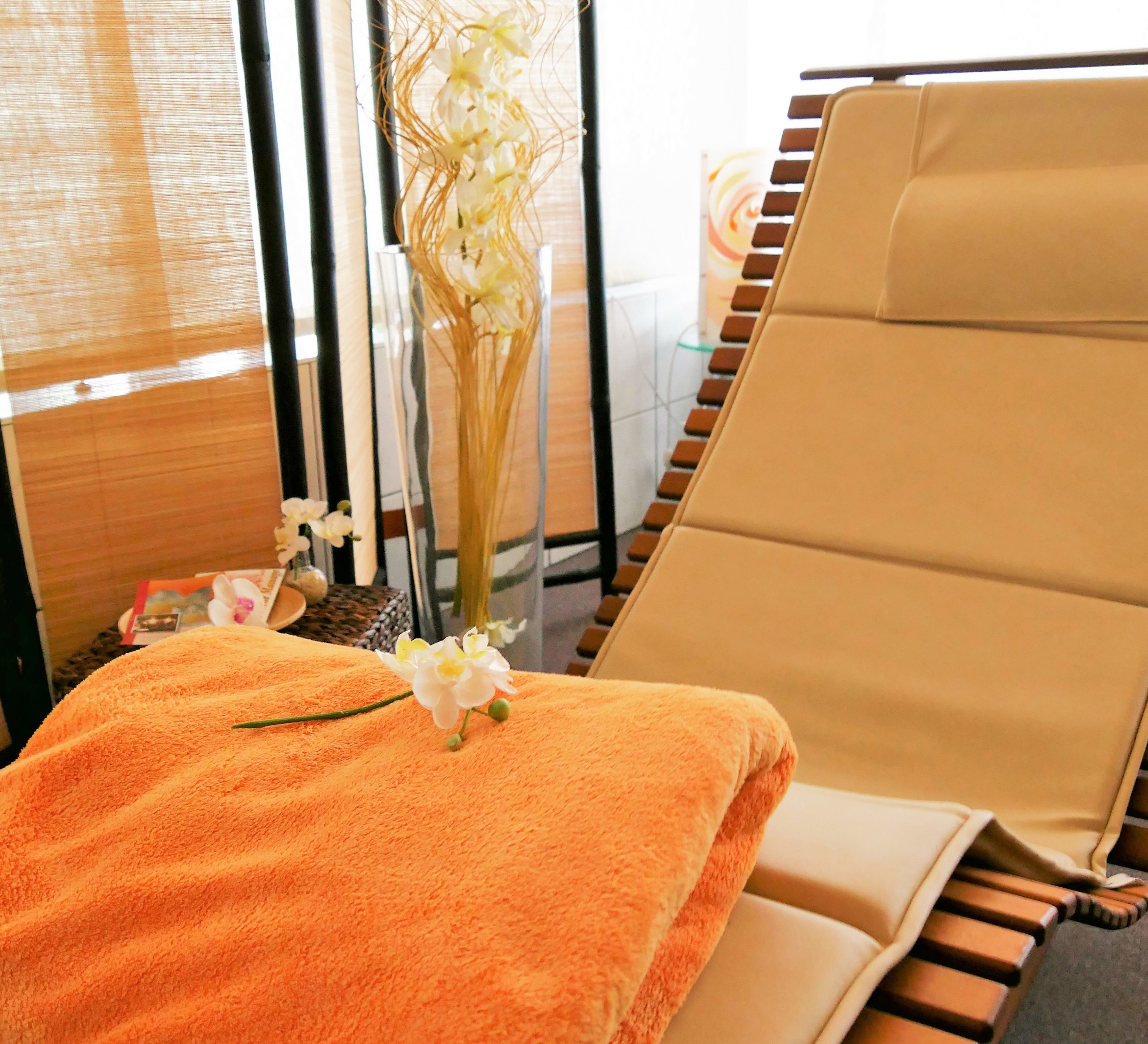 Eine ergonomisch geformte Liege aus Holz mit beigem Polster und einer weichen orangenen Decke. Dahinter ein Paravant und Dekoblumen in einer großen Vase.