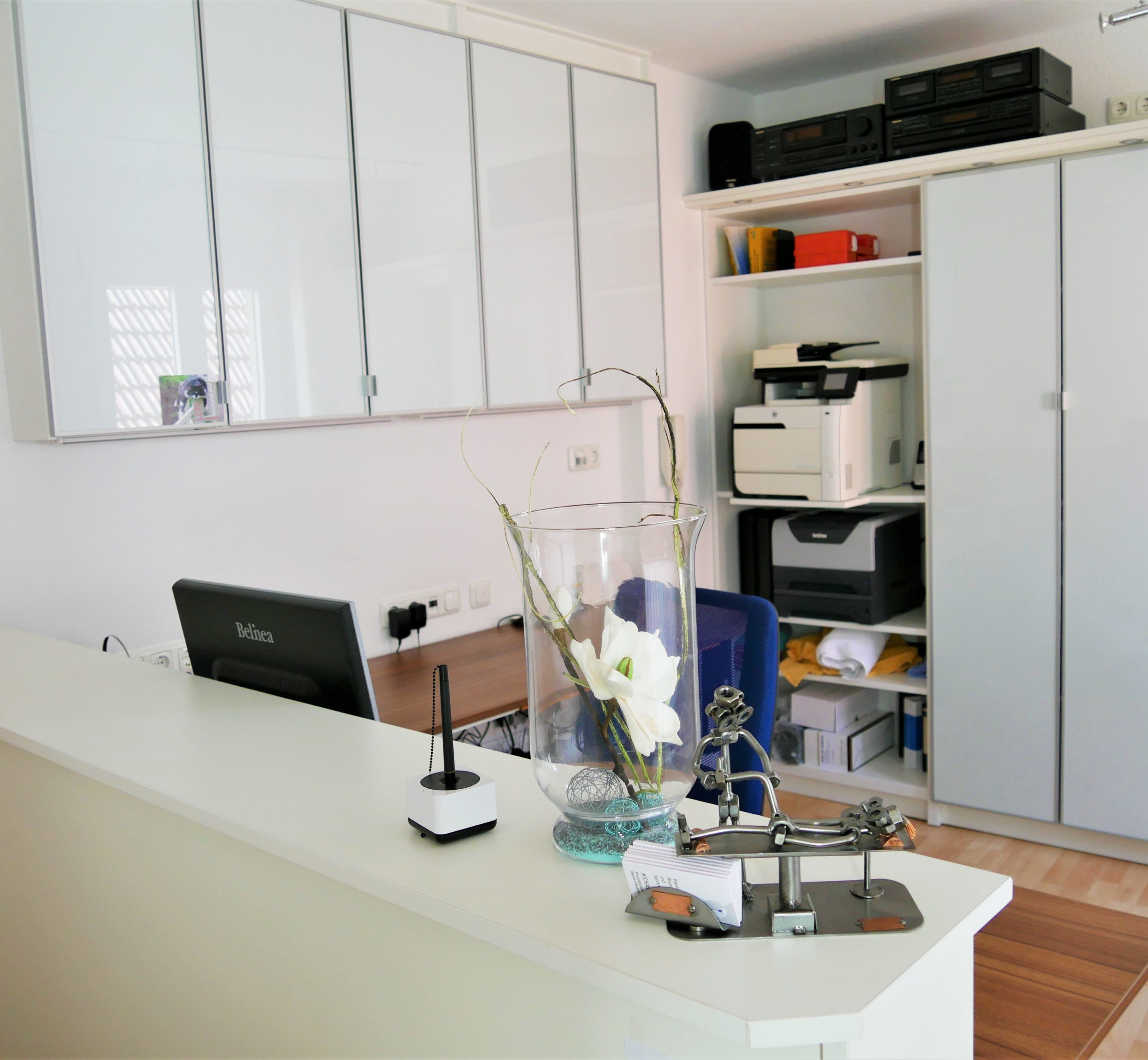 Eine weiße Theke.. Darauf eine Glasvase mit Blume, Dekoration und ein Kugelschreiber. Dahinter ein Holzschreibtisch mit Computer. Im Hintergrund Regale mit Drucker und Ordnern.