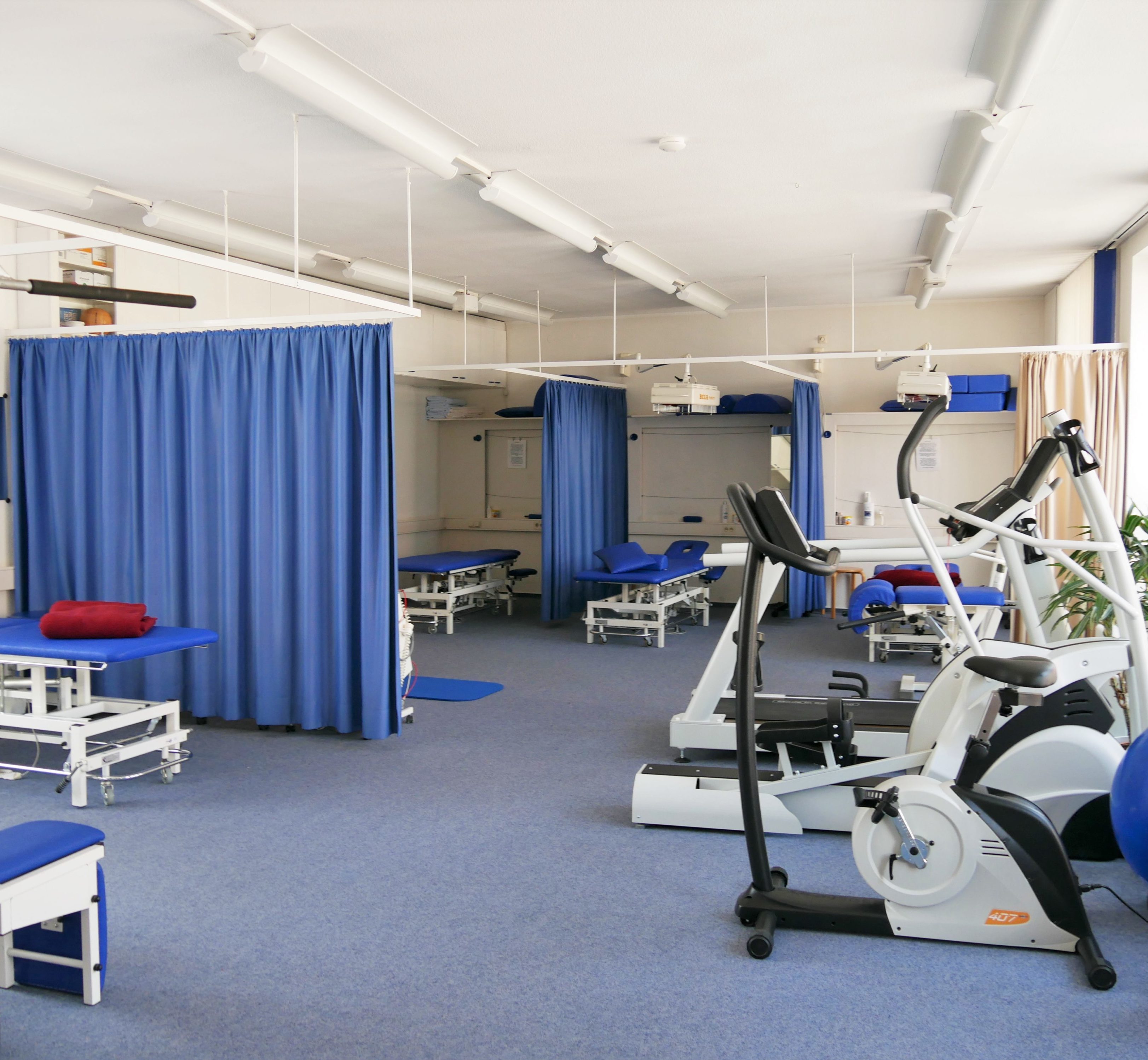 Ein großer Raum mit Liegen, getrennt durch Vorhänge. An der großen Fensterfront rechts mehrere Trainingsgeräte