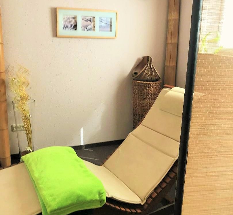 Eine ergonomisch geformte Liege aus Holz mit beigem Polster und grüner Decke hinter einem Paravant. Dahinter verschiedene Dekoartikel wir Bilder und Vasen.